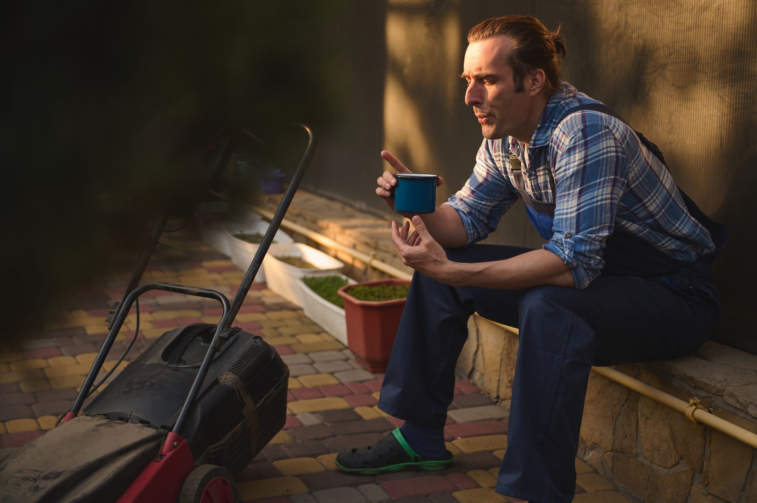 Man wearing gardening overalls and plaid shirt, sitting with a mug of coffee near a lawn mower