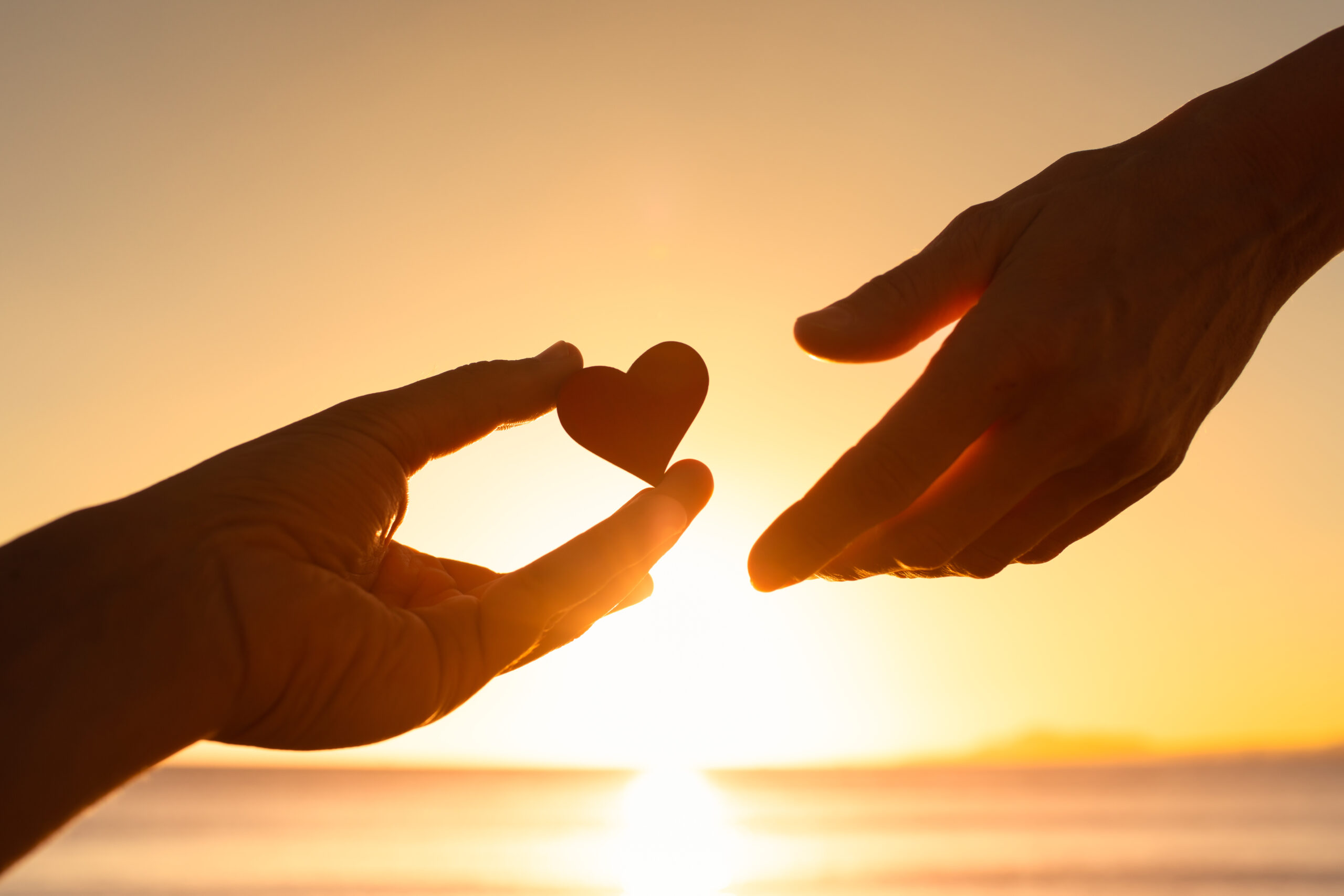 silhouette of one hand passing a heart-shaped token to another hand against the background of a sunrise