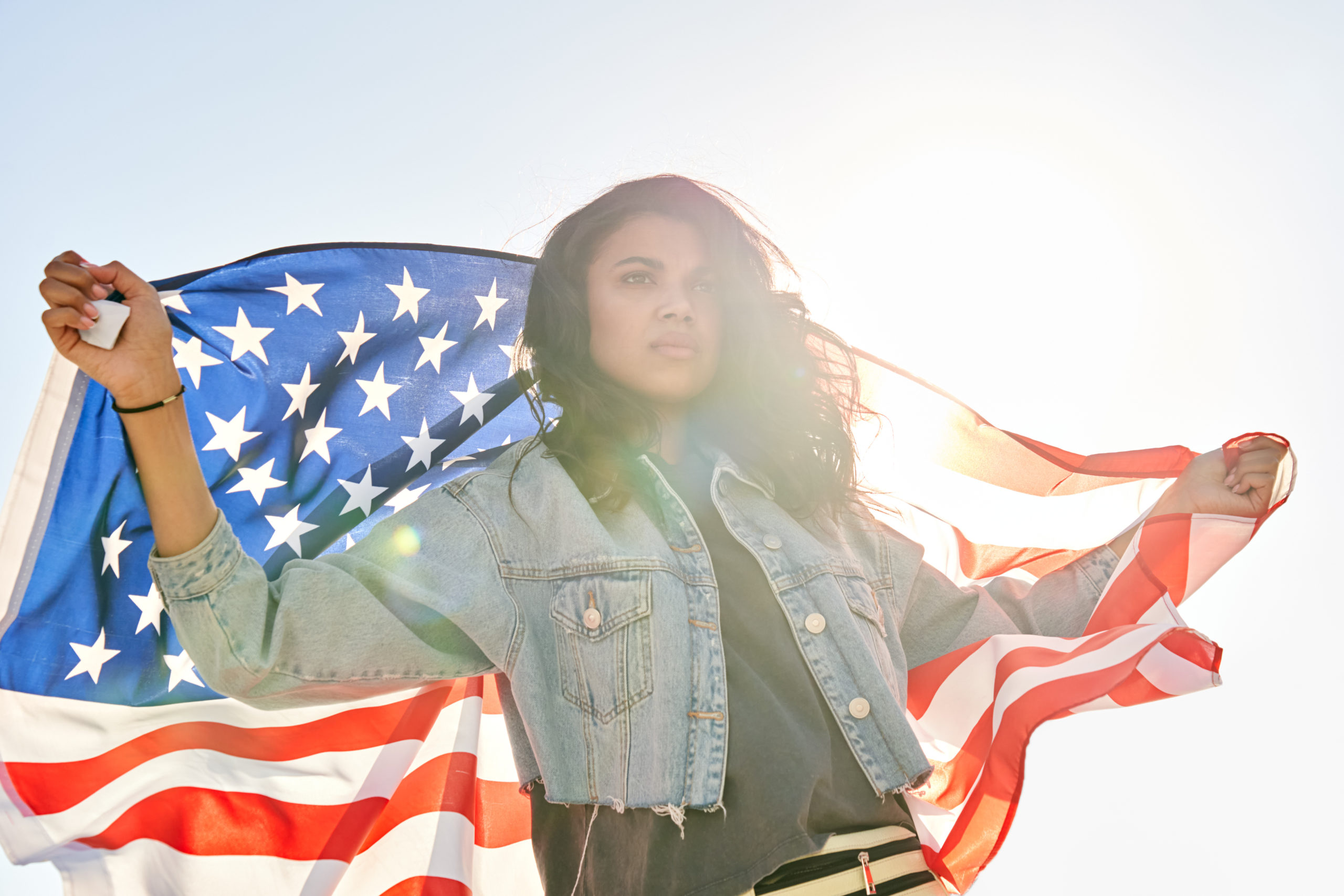Proud serious young Black American woman standing on street holding the American flag looking away backlit with sun.