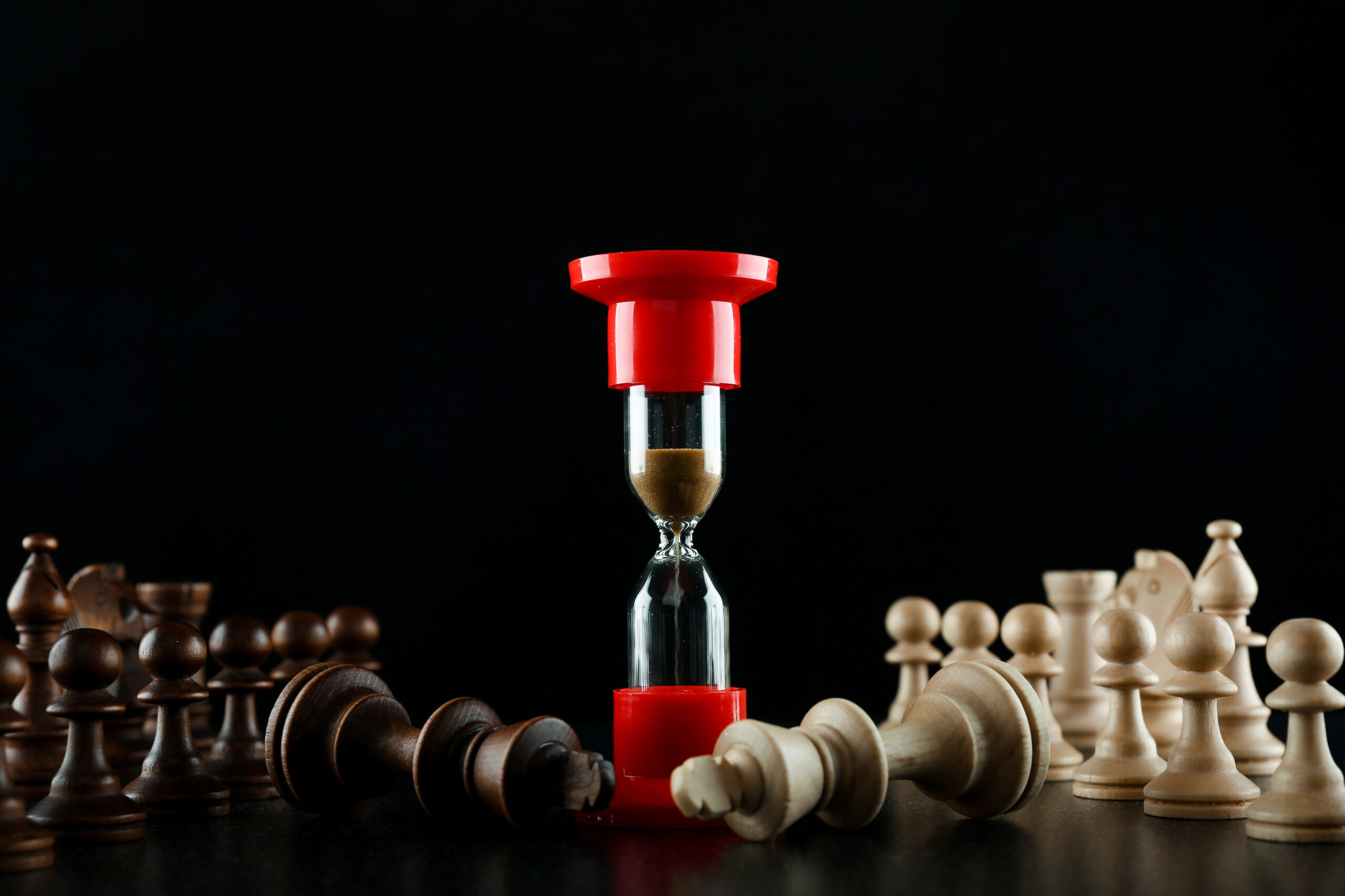 an hourglass on a chessboard next to two fallen king chess pieces