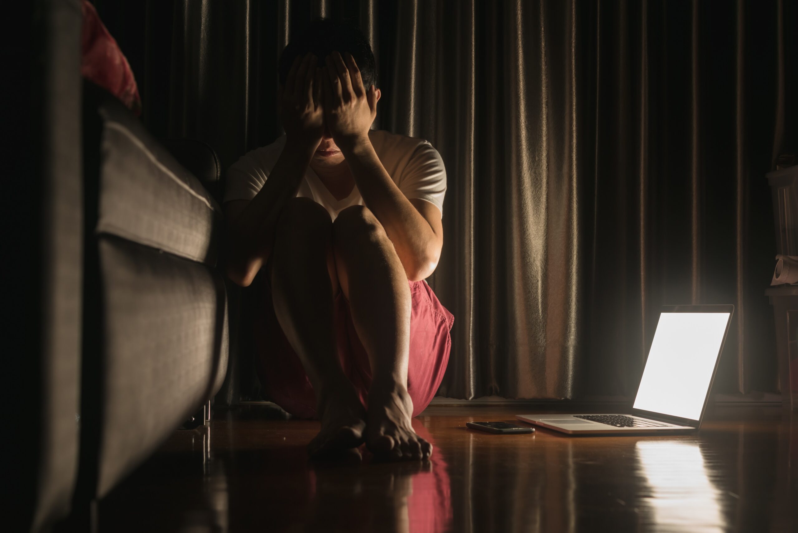 Person with hands on face (sign of depression) sitting on floor with laptop beside them