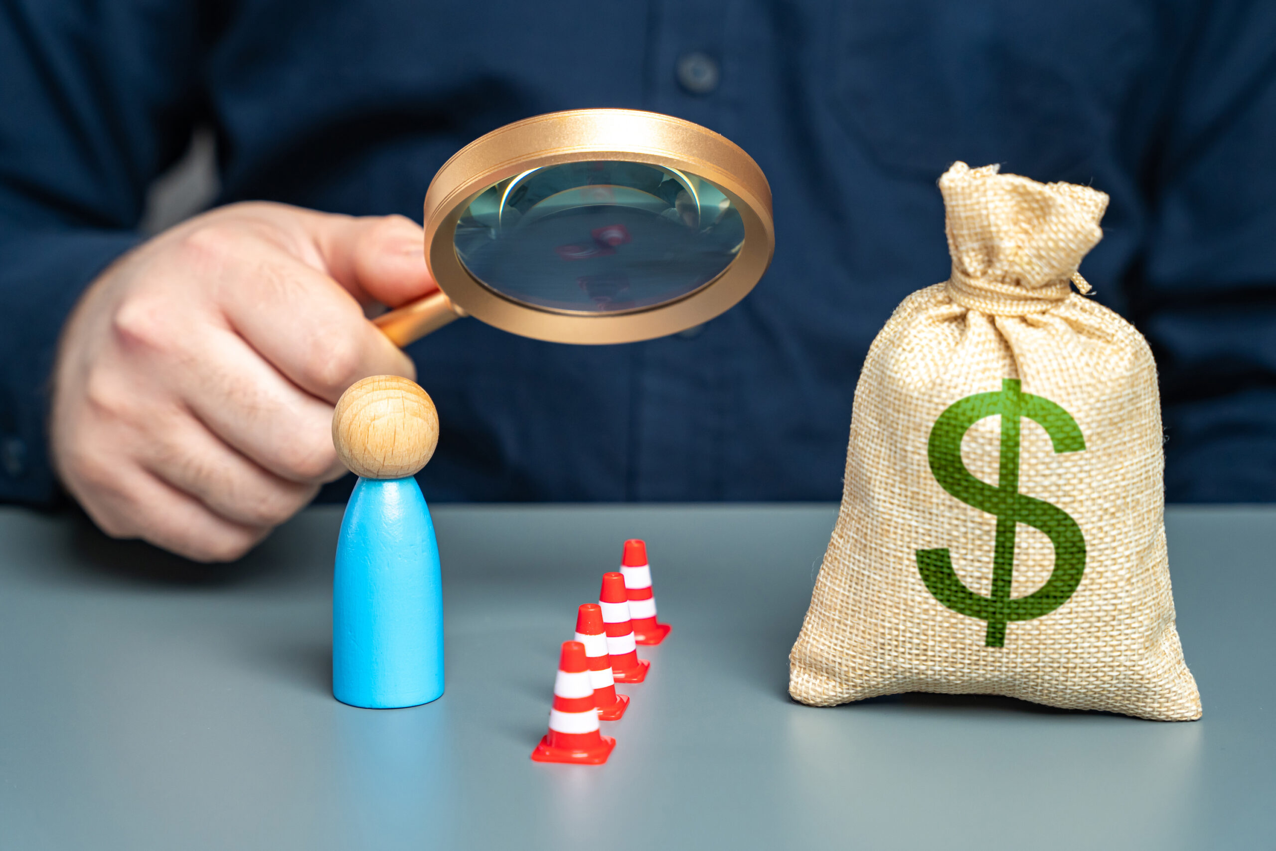 Bag of money with red pylons blocking symbol for person with magnifying glass examining barrier