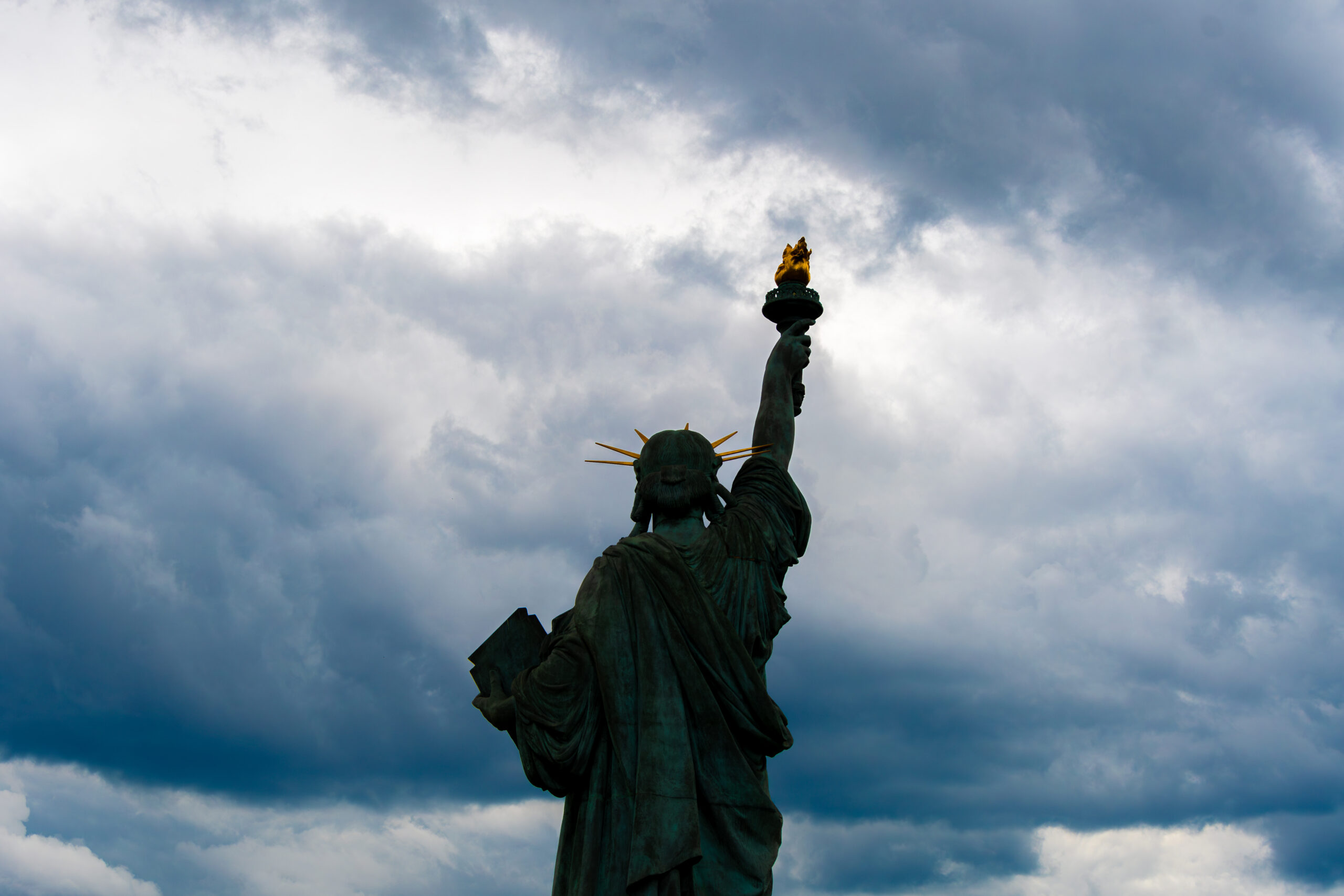 Statue of Liberty in shadows against cloudy sky but with torch still lit