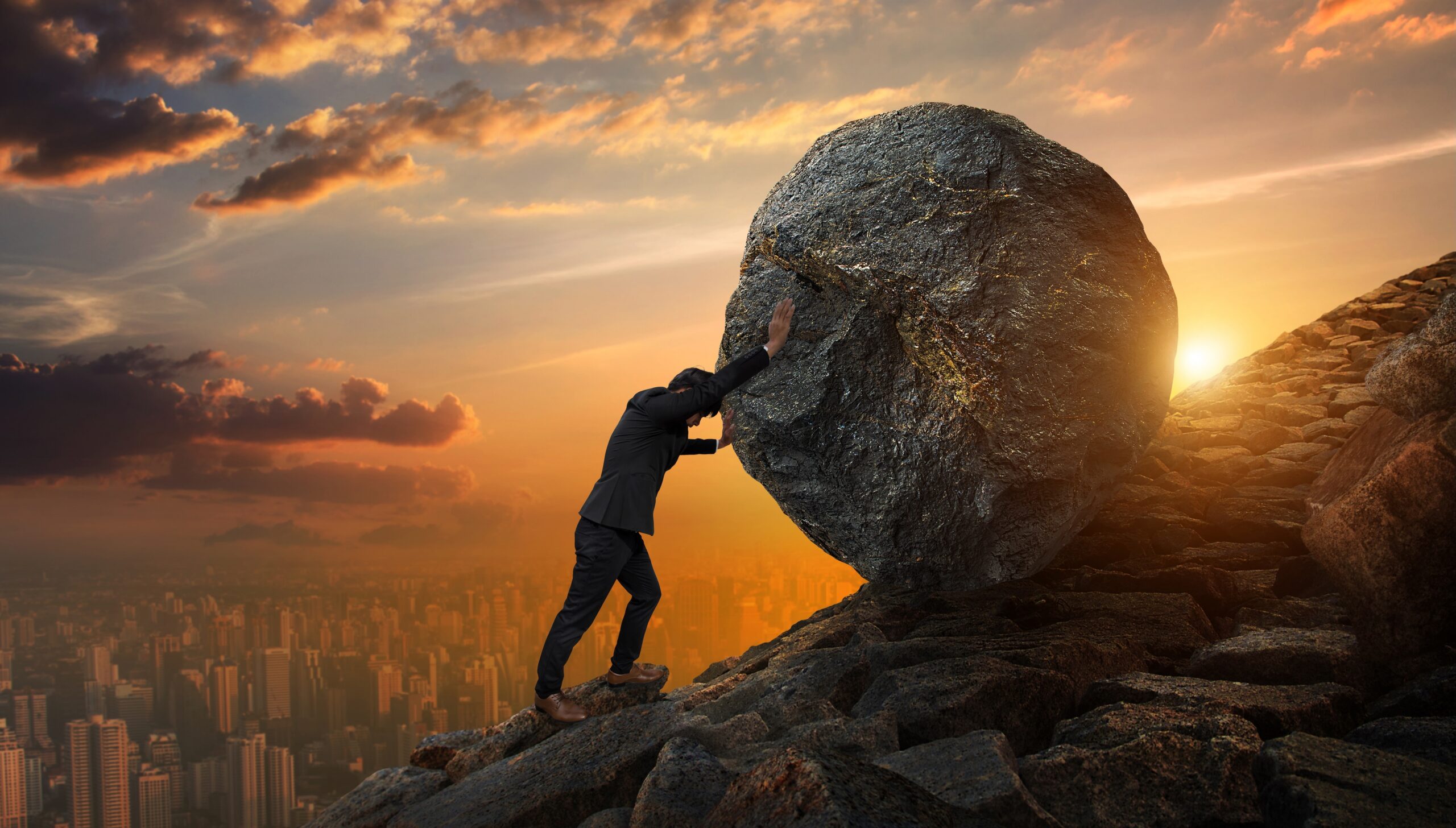 Business man pushing large stone up a hill at sunset