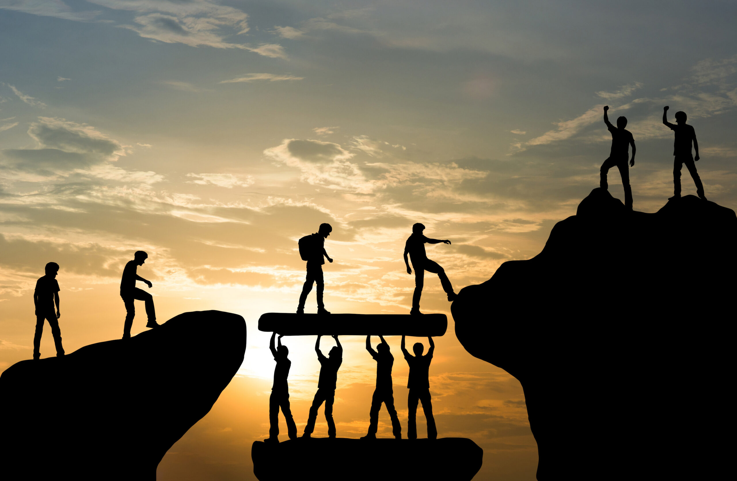 Four persons lifting bridge between ledges allowing people to cross to peak - sunrise background